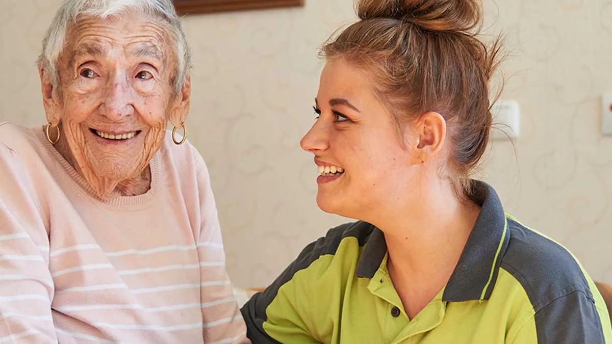 Pflegerin blickt lächelnd zu einer ebenfalls lächelnden Bewohnerin von PFLEGEN & WOHNEN HAMBURG Marienthal.