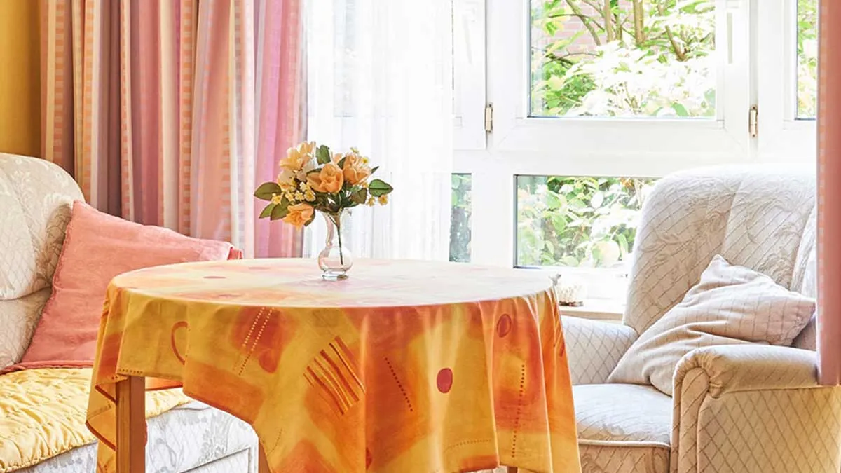 Heller Raum im PFLEGEN & WOHNEN HAMBURG in Horn. Vor einem Fenster stehen ein heller Sessel, ein helles Sofa und ein Tisch mit Blumen und gelber Tischdecke.