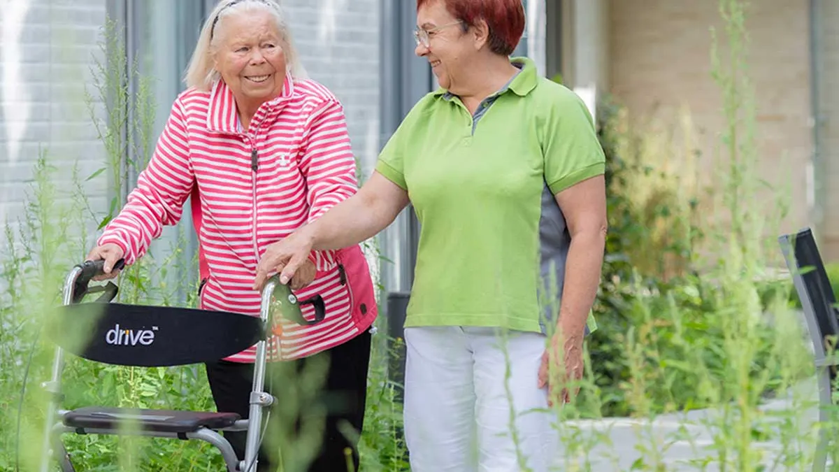 Bewohnerin mit Rollator geht zusammen mit Pflegerin am PFLEGEN & WOHNEN HAMBURG-Standort Marienthal spazieren.