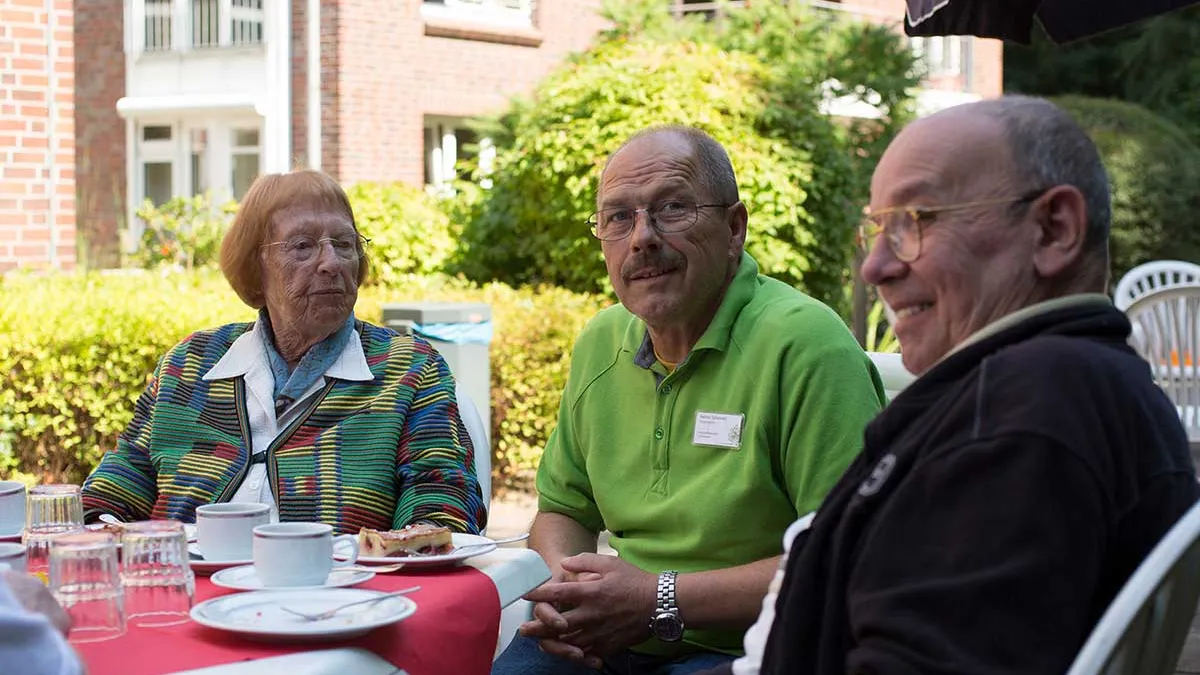 Pfleger sitzt draußen zusammen mit Bewohnern von PFLEGEN & WOHNEN HAMBURG am Standort Lutherpark bei Kaffee und Kuchen.