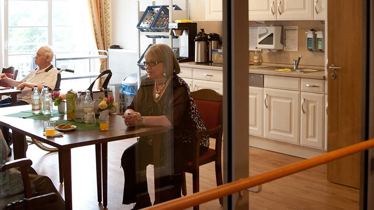 Besucherin unterhält sich mit Bewohner an einem Tisch im PFLEGEN & WOHNEN HAMBURG Holstenhof. Im Hintergrund ist eine kleine Küche.