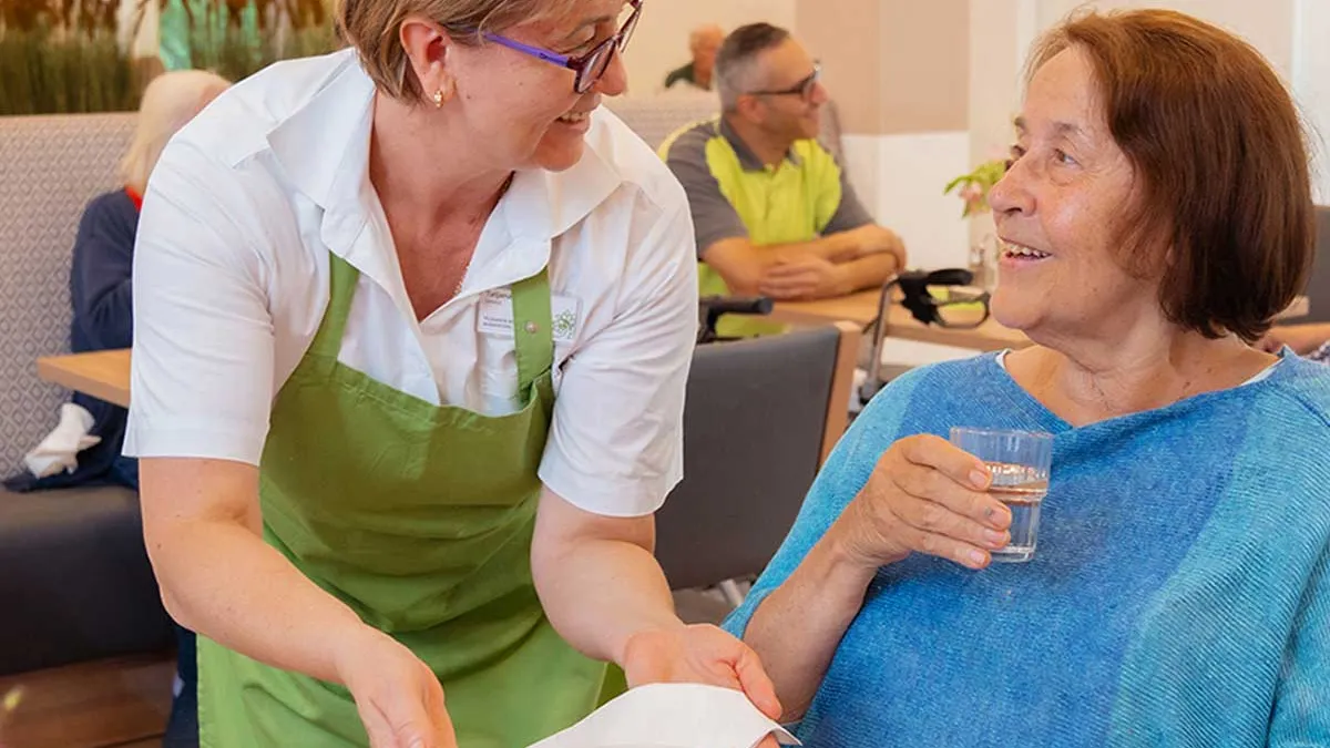 Mitarbeiterin von PFLEGEN & WOHNEN HAMBURG am Standort Marienthal serviert einer sitzenden Bewohnerin Essen.