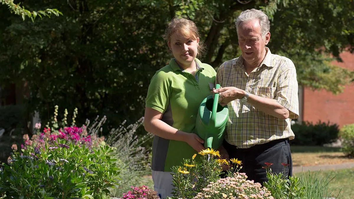 Pflegerin hilft Bewohner von PFLEGEN & WOHNEN HAMBURG Lutherpark beim Gießen von Blumen.