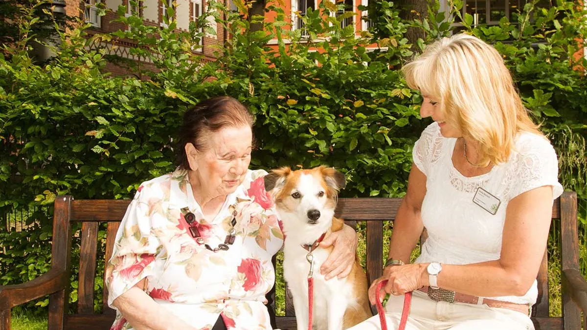 Bewohnerin im PFLEGEN & WOHNEN HAMBURG Farmsen sitzt mit einem Hund und einer Mitarbeiterin auf einer Bank.