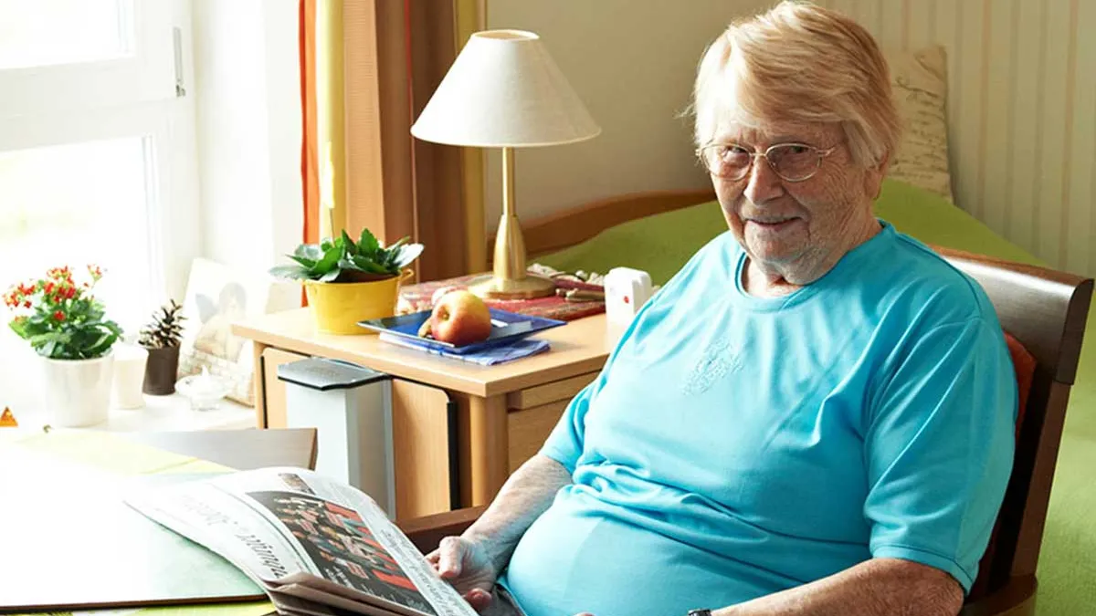 Bewohnerin sitzt am Tisch und liest Zeitung in ihrem Raum im PFLEGEN & WOHNEN HAMBURG Holstenhof. Hinter ihr stehen ein Bett und ein Nachttisch.