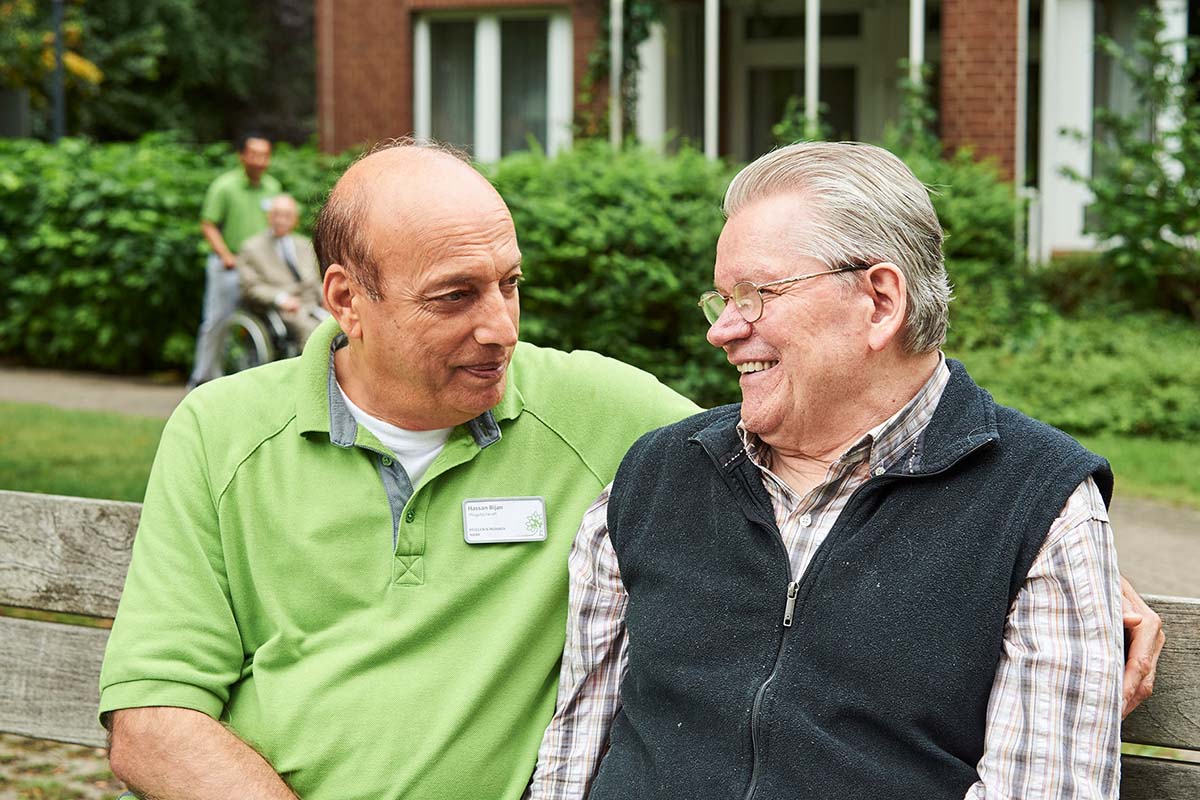 Ein Bewohner und Mitarbeiter auf einer Parkbank im Grünen vor dem PFLEGEN & WOHNEN HAMBURG Horn Gebäude, in zentraler Lage.
