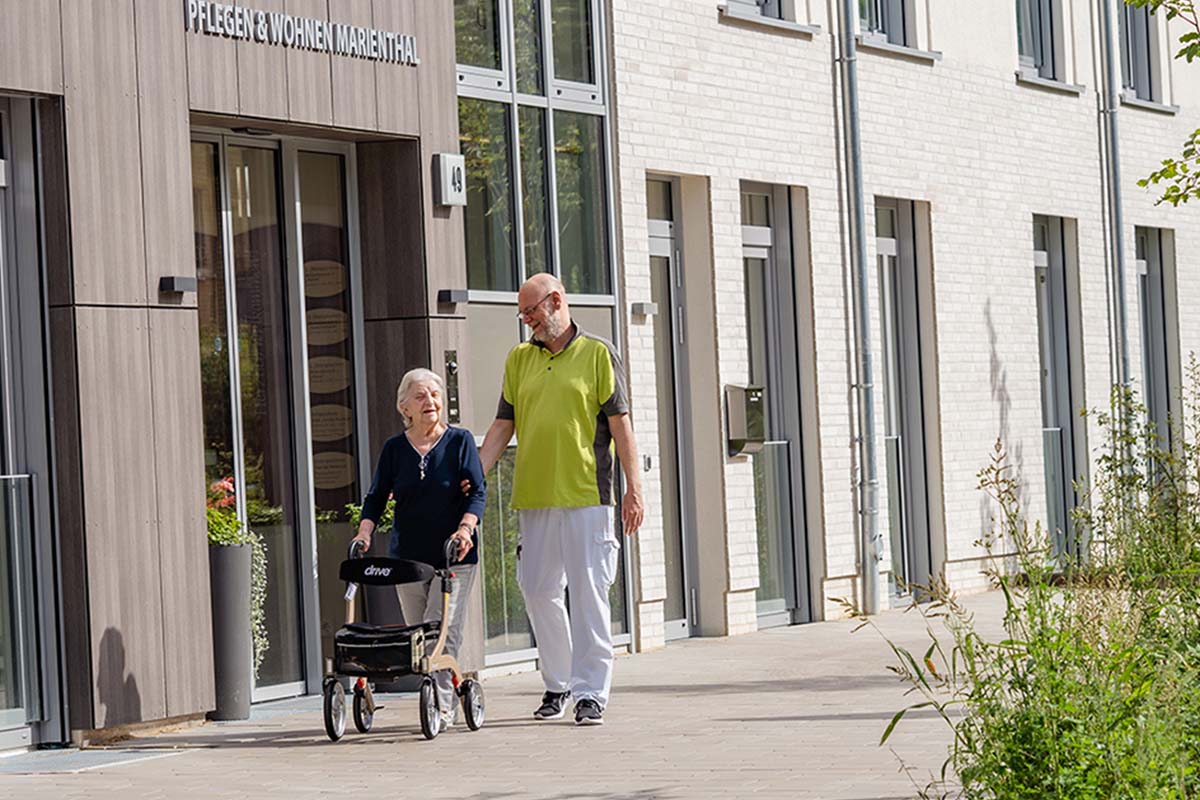 Ein Pfleger bei PFLEGEN & WOHNEN HAMBURG Marienthal bei einem Spaziergang mit einer Bewohnerin mit Gehhilfe bei Sonnenschein.