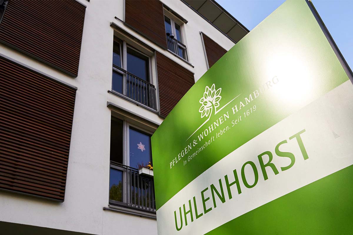 Detailansicht: grünes Eingangsschild von PFLEGEN & WOHNEN HAMBURG Uhlenhorst mit modernem Gebäudekomplex für Seniorenpflege.